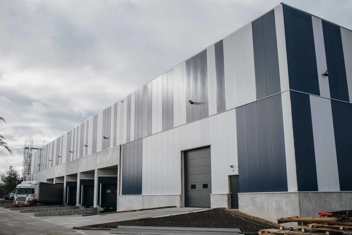 De buitenkant van de nieuwe productiehal. Het is een klassiek industrieel gebouw met lichtgrijze en donkergrijze/blauwe panelen.