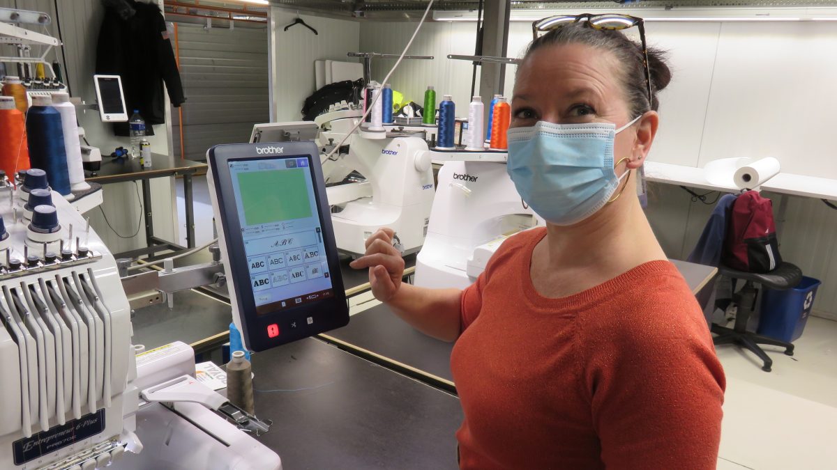Een collega van Bewel demonstreert hoe een van de stikmachines in de Bewel stikkerij werkt. Ze draagt een mondmasker.
