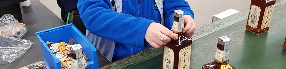 Een medewerker van Bewel bevestig een label op een fles van Jack Daniels. Enkel zijn handen zijn in beeld.