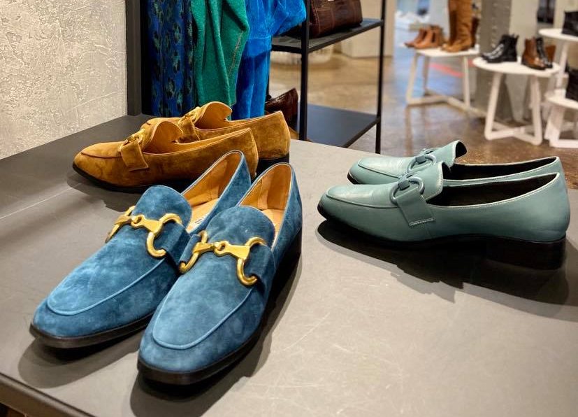 Drie paar schoenen van La Bottega staan netjes opgesteld in hun winkel. Een paar is bruin, een ander blauw met een gouden gesp en de andere grijsgroen.