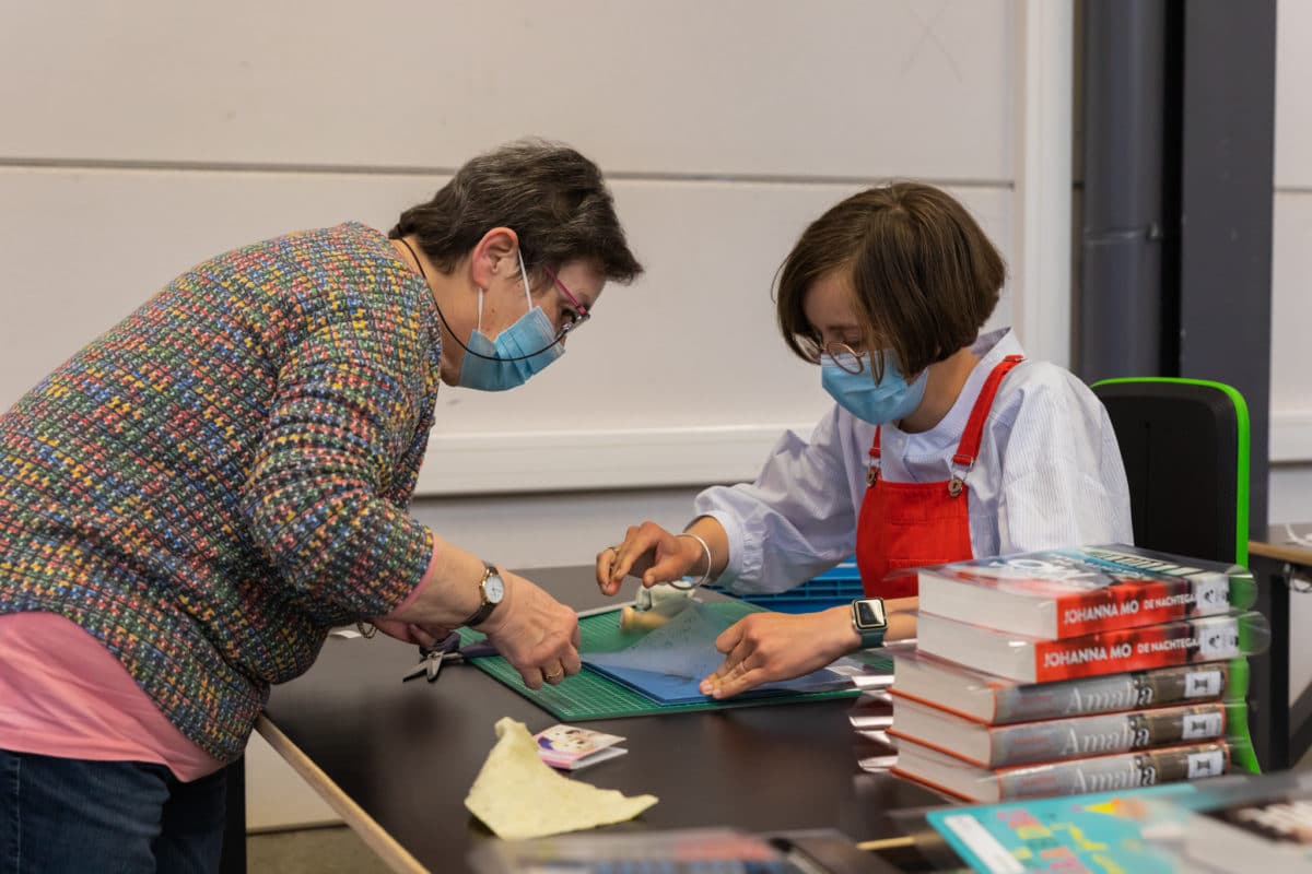 Twee medewerkers van Bewel Hasselt zijn bezig met boeken te kaften. De ene medewerker helpt de andere.